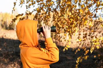 Young woman in yellow sweatshirt and hood with binoculars watching birds in the autumn forest...