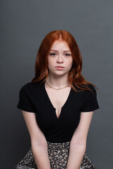 Beautiful teenage young lady with long red hair sitting posing with simple plain neutral gray or...