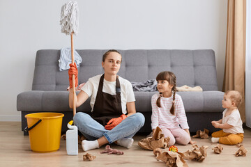 Young adult female sitting on floor among mess with her children, woman holding mop, doing domestic...