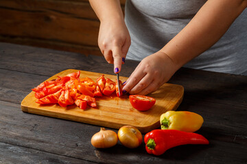 A woman in the kitchen with a knife cuts red tomatoes on a wooden board on the table next to onions and peppers.