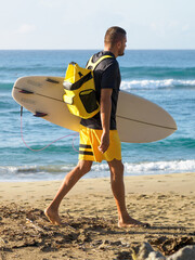 Surfer in yellow shorts and backpack walking on beach.