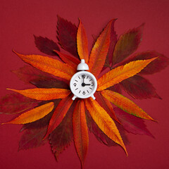 Fall Back, Autumn Time Change Concept. White Alarm Clock and Orange Autumn Leaves on Red Background.
