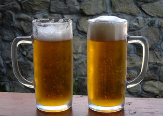 Two glasses of cold draft beer with lush foam on a wooden desk with a stone wall background
