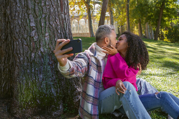 happy couple sitting on the grass in the park kissing and taking a picture with the phone,
