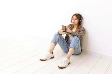 A girl in headphones sits on a white floor in jeans and a shirt and looks at the phone screen. High quality photo