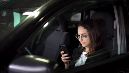 A young businesswoman in a parking lot sits in a car and texts on the phone. A young girl in glasses and a suit with a phone in her hands.