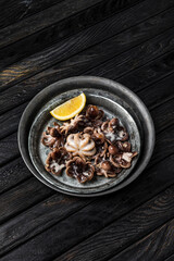 A dish of cooked baby octopus with a slice of lemon, shot from above, dark wooden backdrop