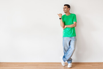 Positive young arab guy pointing aside at copy space, standing against white wall, full length portrait, place for ad © Prostock-studio