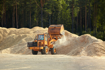 working bulldozer unloads against the background of large mountains of small wooden sawdust - obrazy, fototapety, plakaty