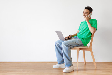 Positive arab man sitting on chair with computer on his lap, having phone call with employer, copy space