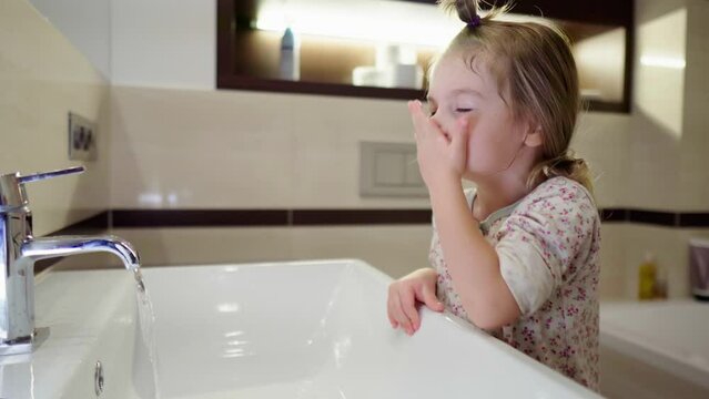 Little girl washes her face with cool water in the bathroom. Children's daily hygienic water procedures. The child finishes washing and turns off the tap. Formation of healthy habits from an early age