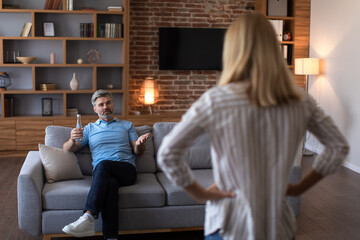 Angry mature caucasian wife scolds at husband with bottle of beer on sofa in living room interior