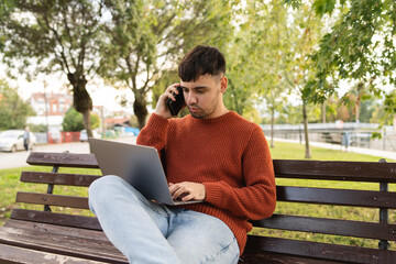 Fototapeta premium A young businessman outdoors in park sitting with his laptop talking on phone enjoying outside 