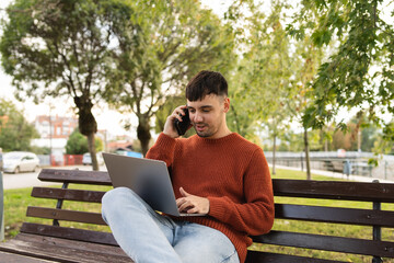 A young businessman outdoors in park sitting with his laptop talking on phone enjoying outside 