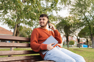 A young businessman outdoors in park sitting with his laptop talking on phone enjoying outside 