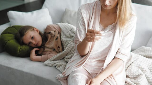 Worried parent call doctor nervous of child fever. Mom looking at thermometer and sick child daughter lying on background. Caring mum treating sick kid at home. Family healthcare and health concept