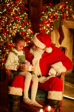 Vertical Portrait Of Cute African American Girl Sitting In Santas Lap And Holding Christmas Present In Cozy Setting