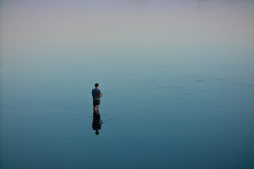 Fototapeta premium A fisherman stands in the water and catches fish with a fishing rod. River or lake, no waves