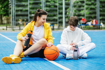 Teenager boy and girl are sitting on sports court, drinking water and resting during break.