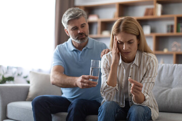 Worrying mature caucasian husband calms wife, suffering from headache, gives glass of water on sofa