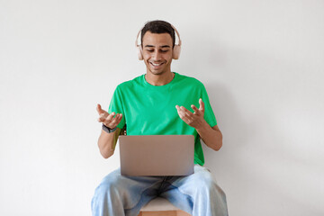 Excited arab man having video conference on laptop, wearing headphones and talking at webcamera, sitting on chair