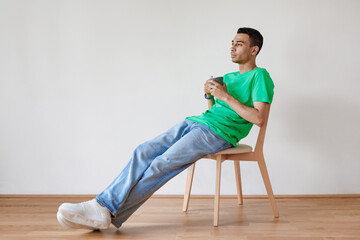 Rest concept. Pensive arab guy drinking coffee, sitting on chair over white wall, free space, full length shot