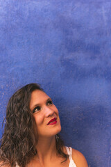 Image vertical of a brunette woman looking up while grimacing with a smile leaning against a blue wall inside the narrow colorful streets of Villajoyosa, Alicante, Spain.w