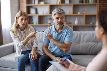 Sad mature caucasian lady and male point fingers at each other in clinic office interior at meeting