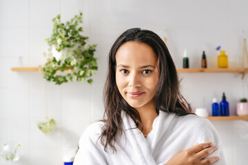 Portrait of young beautiful woman with dark skin in white bathrobe with cosmetic bottle on the background. Confident young woman feeling harmonous and calm in her body. Wellness and body positivity