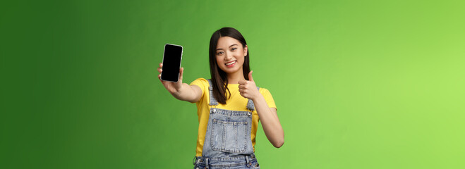 Cheerful cute asian woman judging good smartphone game, show thumb-up like sign, extend arm hold...