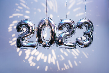 Silber foil balloons in numbers shape 2023 hanging against blue wall with fireworks projection. New...