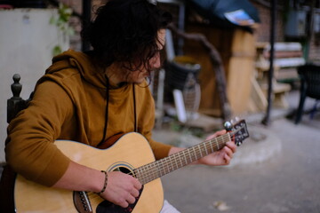 One Asian guy with medium length black curly hair sits outside on a wooden chair and plays the guitar.