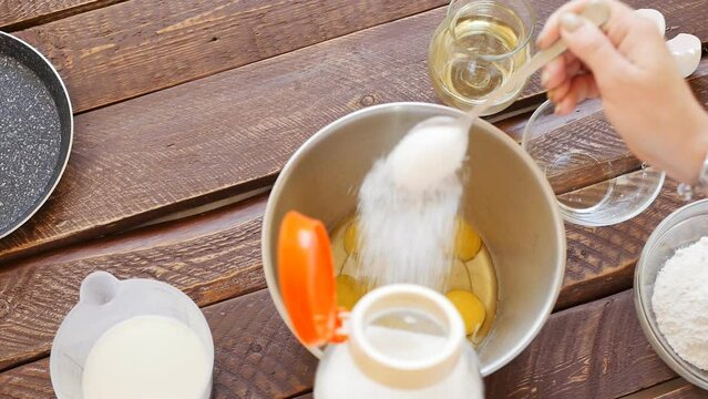 Top view woman pours granulated sugar with a spoon into a bowl with egg yolks in the process of preparing dough for pancakes on a brown wooden table from boards