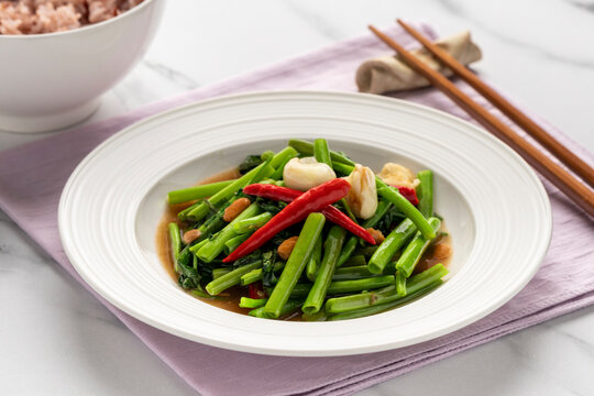 Stir fired Chinese water morning glory or water spinach with soybean paste, a simple Thai-Chinese dish named pad pak boong, served on a white serving plate together with a bowl of brown jasmine rice.