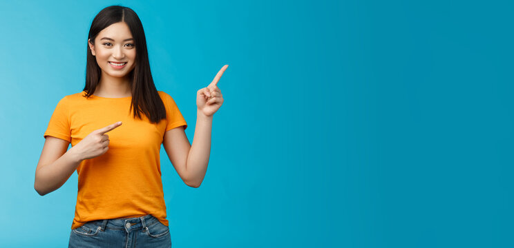 Friendly cheerful asian brunette girl inviting visit link interesting place, pointing upper right corner smiling joyfully camera, telling you about good promotion, stand blue background upbeat