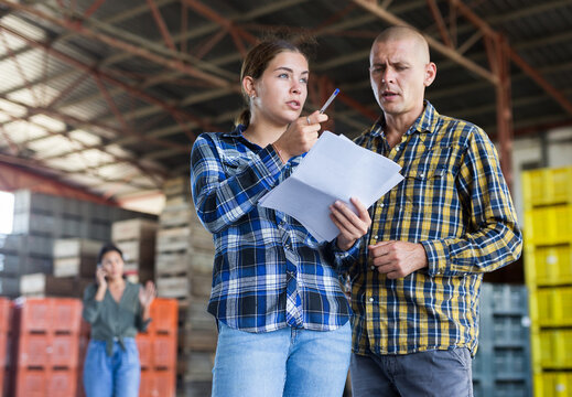 Man and woman discussing details of agreement while standing in warehouse. Another woman talking on phone behind them.