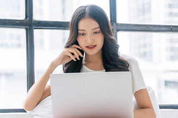 Fototapeta premium Happy asian casual woman reading news writing online with laptop or shopping comfortable at home. Freelance young girl using notebook relax, work sitting with pillow beside window.Lifestyle Technology
