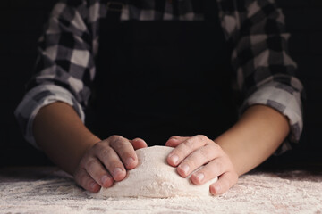 Man kneading dough at wooden table on dark background, closeup