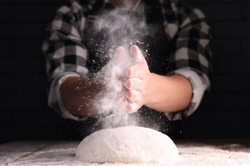 Man sprinkling flour over dough at wooden table on dark background, closeup
