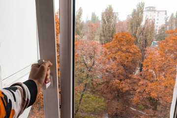 The man's hand opens a white plastic window with autumn trees behind the window. Dark day. Heating....