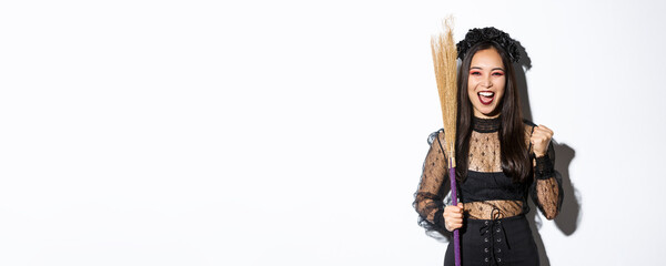 Image of cheerful asian girl in witch costume celebrating victory, holding broom, saying yes and raising fist in triumph, white background