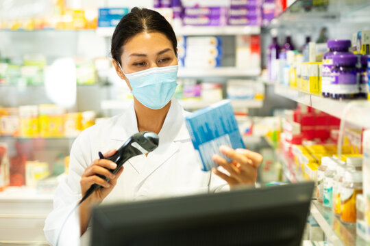Portrait of female pharmacist in medical mask working at the cash register in pharmacy - scans barcode on the medicine package