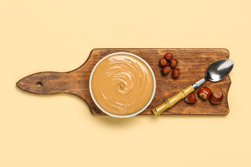 Wooden board with bowl of hazelnut butter on color background