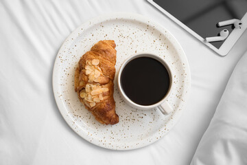 Plate with delicious croissant and cup of aromatic coffee on bed