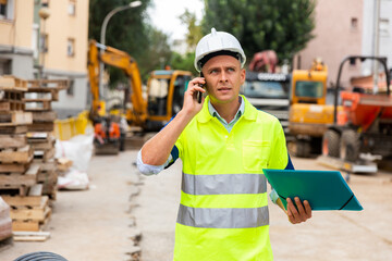 Young confident engineer working at a construction site is talking on a mobile phone, discussing important issues