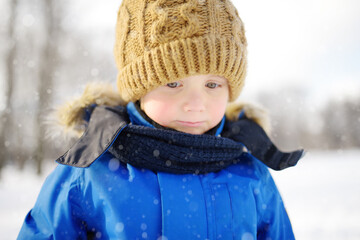 Fototapeta na wymiar Portrait of little upset boy during walk in snowy winter park. Kid dressed in warm clothes, hat and scarf. Active winter outdoors leisure for child