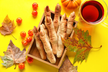 Box with Halloween witch finger cookies, candies, leaves and glass cup of tea on yellow background