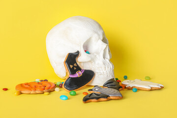 Tasty Halloween cookies with candies and human skull on yellow background