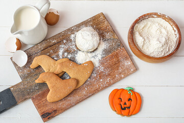 Board with Halloween cookies, raw dough, flour and jug of milk on white wooden background