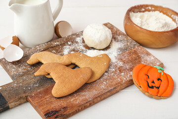 Board with Halloween cookies, raw dough, egg shells and jug of milk on white wooden background